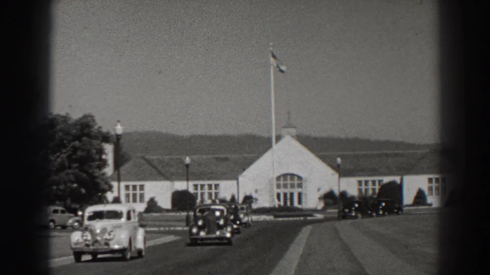 1938 8mm view at Bonneville Dam, Oregon, with vintage cars on a paved road before a white gabled building and flagpole.