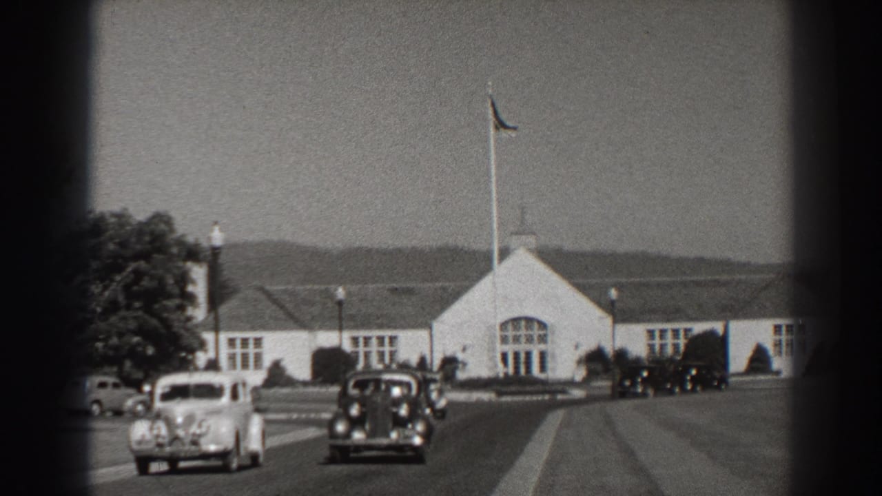 Light and dark period cars pass in front of a white building with arched windows.