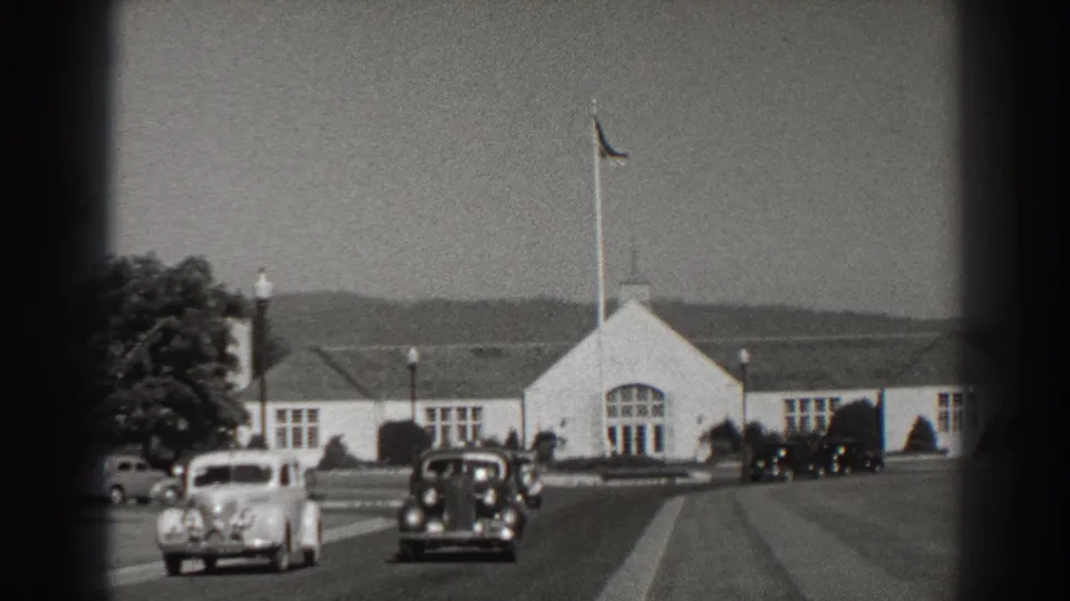 Vintage automobiles pass the white Bonneville Dam building, with a tall flagpole, lawns, and distant hills visible.