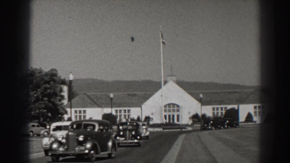 A dark 1930s automobile crosses the foreground at Bonneville Dam, with more cars and the white building beyond.