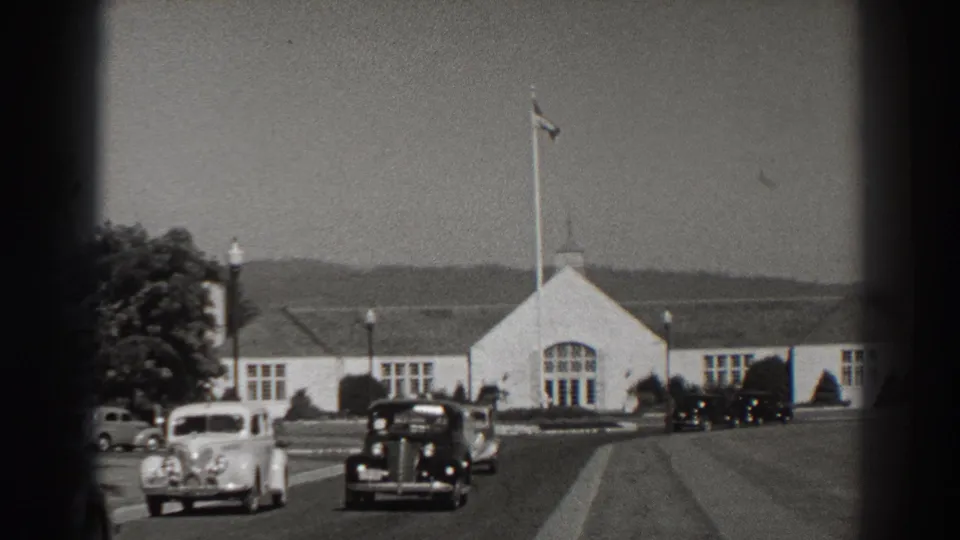 Late-1930s cars move through the Bonneville Dam grounds beside a white building, flagpole, roadway, and landscaped lawn.