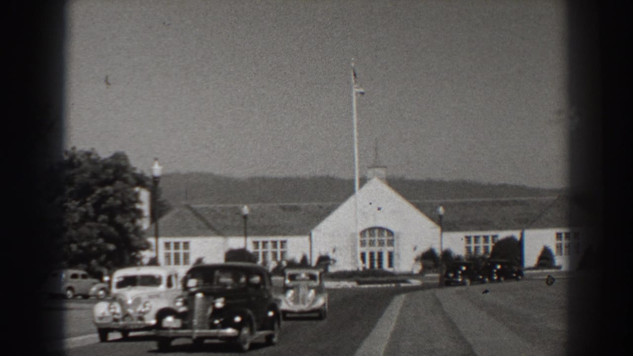 1938 Bonneville Dam-area view with vintage cars on a paved road, a white building, flagpole, trees, and distant hills.