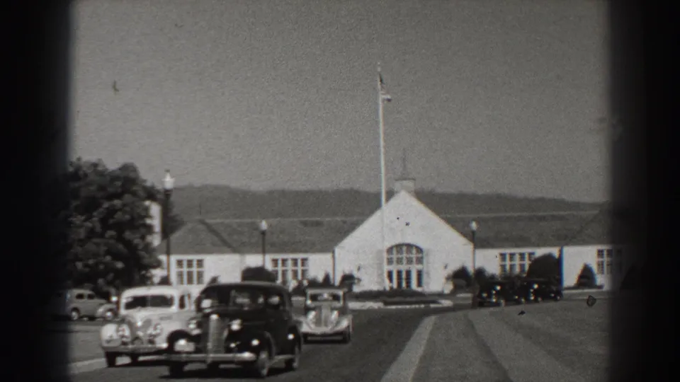 1938 Bonneville Dam-area view with vintage cars on a paved road, a white building, flagpole, trees, and distant hills.