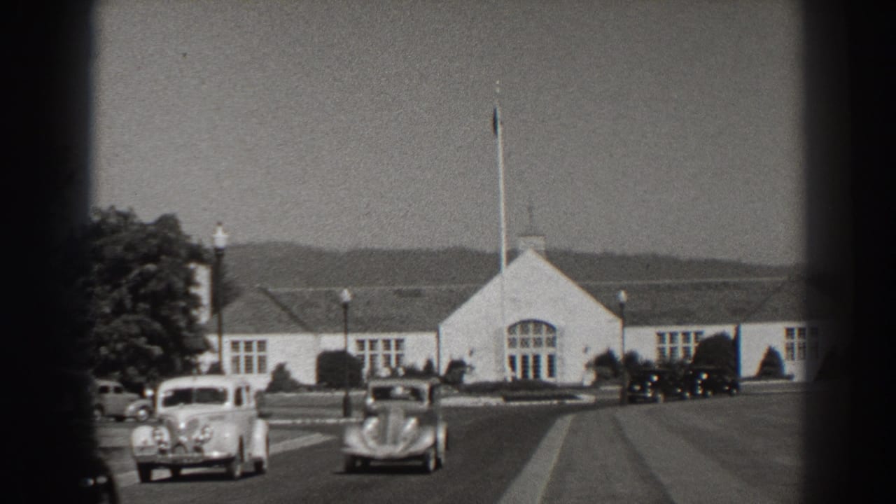 Vintage cars pass in front of a white building with a flagpole and distant hills.