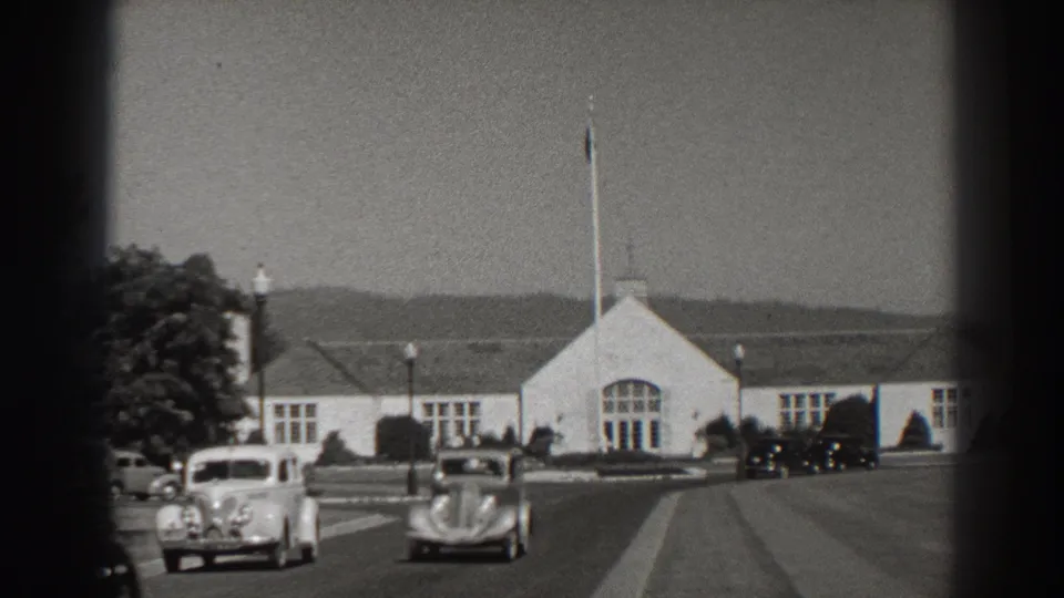 Bonneville Dam-area road scene from 1938 showing classic cars, a white building, a flagpole, trees, and hazy hills.