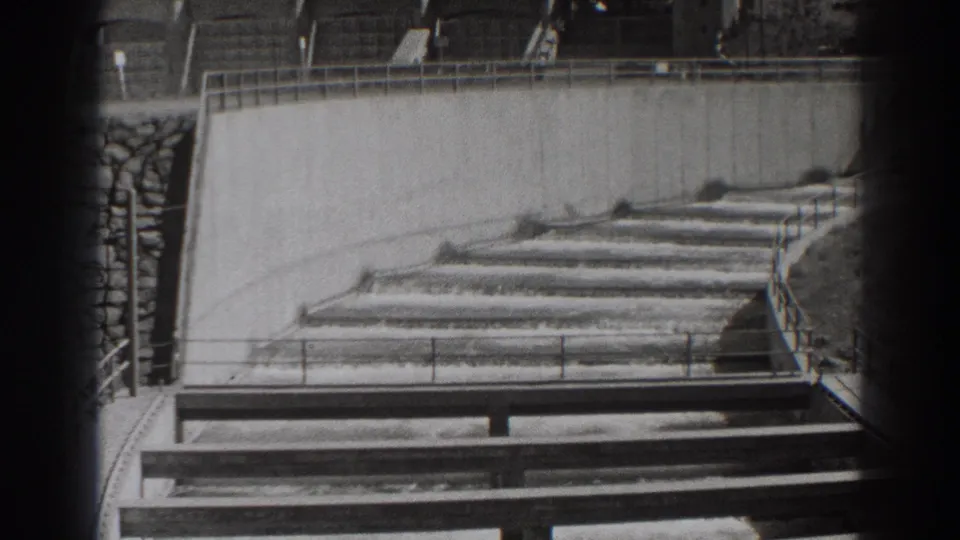 Black-and-white 8mm frame of Bonneville Dam fish-ladder water flowing over stepped weirs below concrete walls.