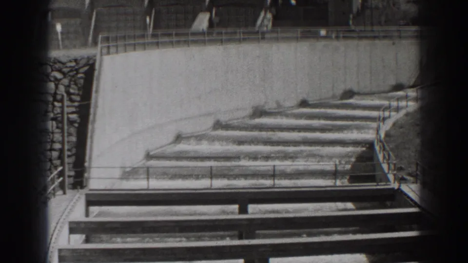 Black-and-white 8mm frame of Bonneville Dam water cascading over stepped concrete sections beneath a railing.