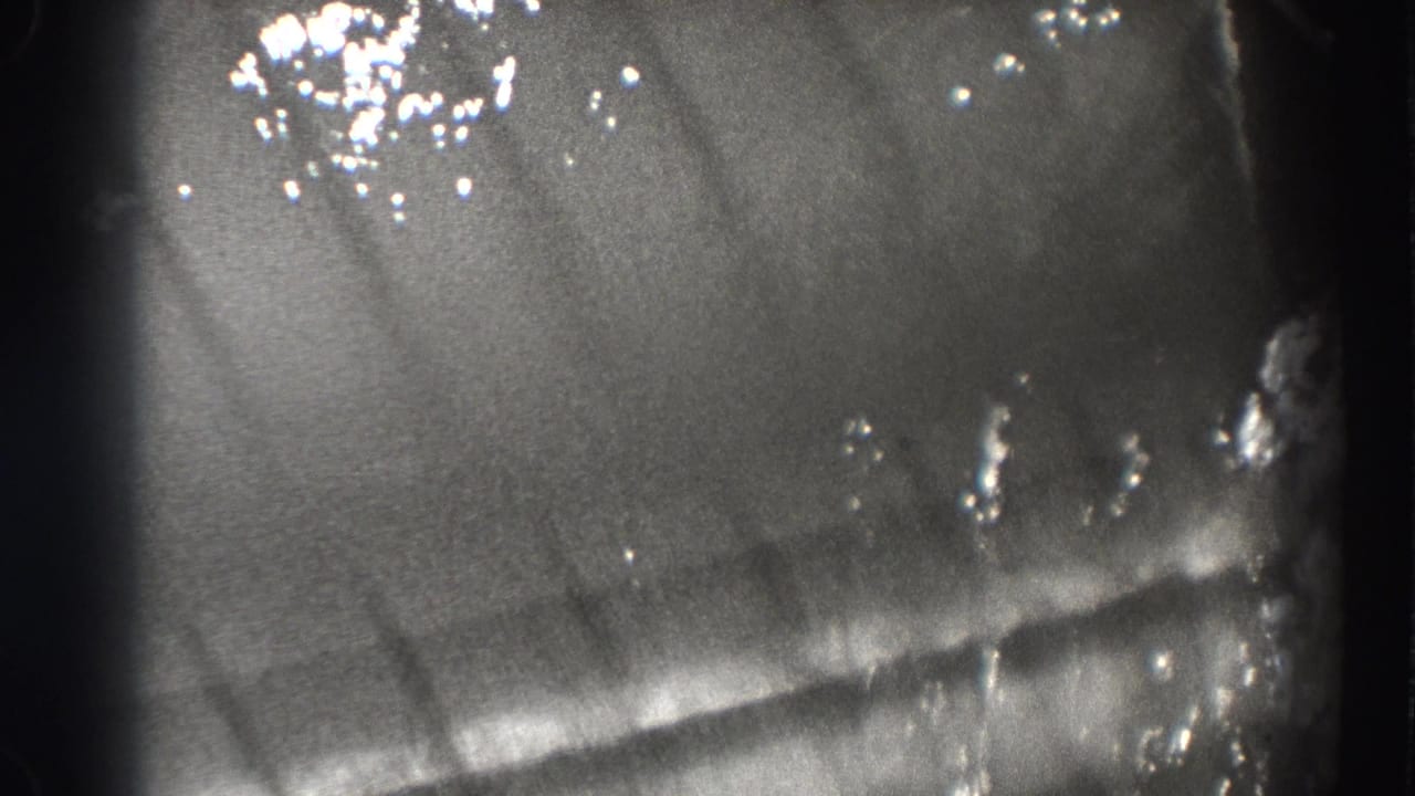 Grainy black-and-white close view of rippling water with bright reflections and faint fish shapes below.