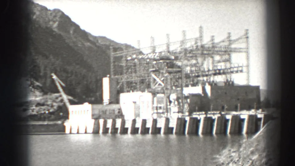 1938 Bonneville Dam, Oregon view across the river toward concrete dam works, steel power structures, and a forested mountain backdrop.
