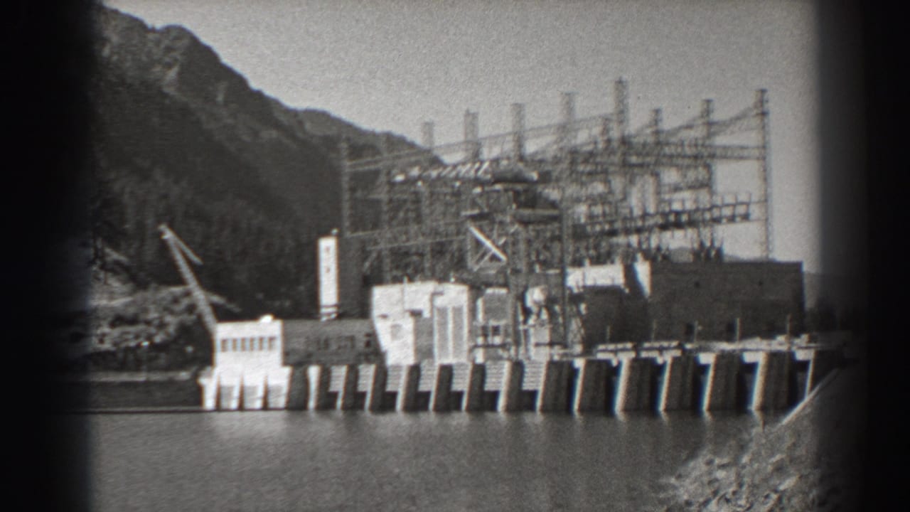 1938 8mm view of Bonneville Dam in Oregon, showing the concrete dam, river, crane, electrical towers, and steep wooded slope.
