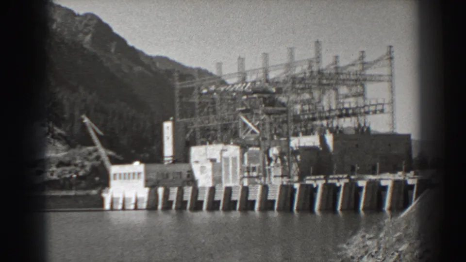 1938 8mm view of Bonneville Dam in Oregon, showing the concrete dam, river, crane, electrical towers, and steep wooded slope.