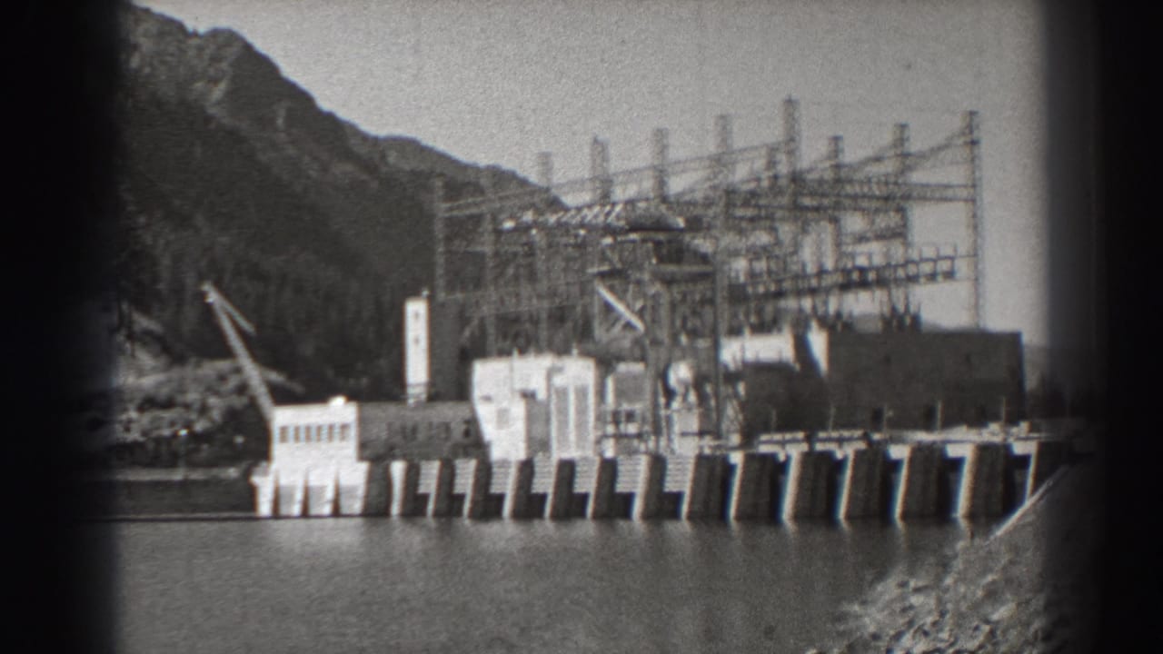 Dam and electrical tower structures viewed across water with mountains in the background.