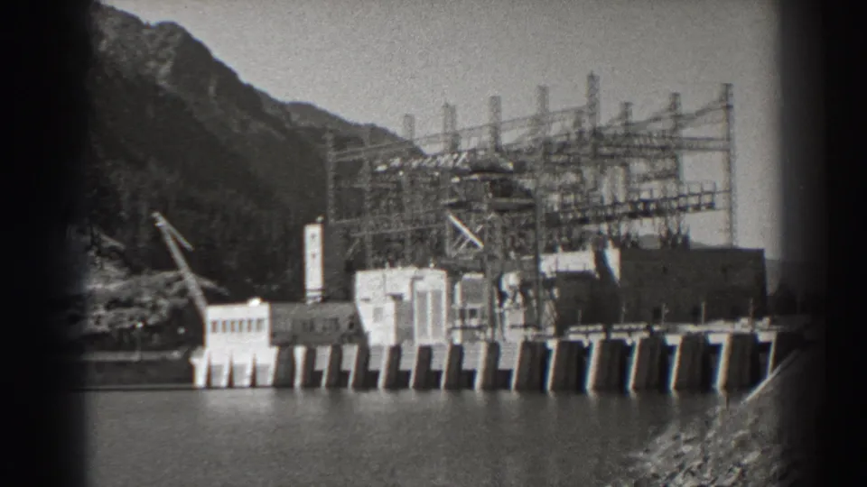 Archival 1938 view of Bonneville Dam with visible spillway gates, a crane, transmission framework, water, and steep Oregon hillside.