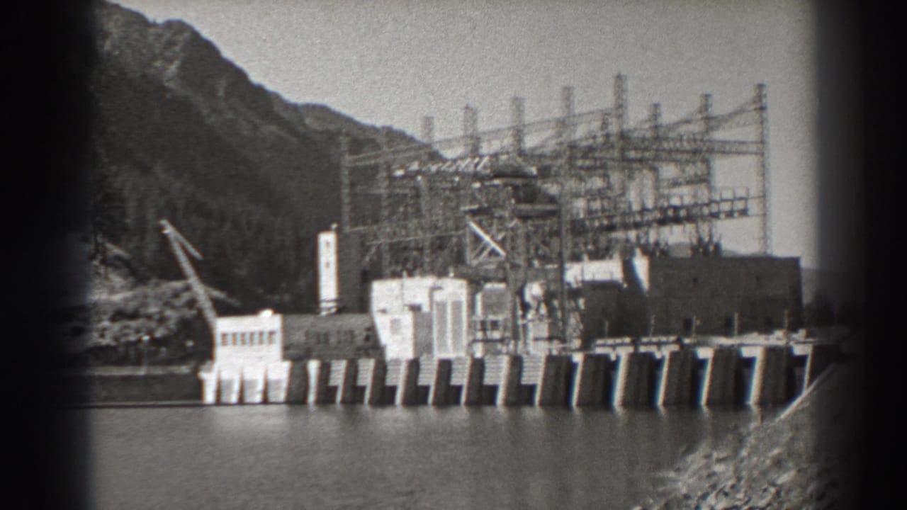 Bonneville Dam construction works beside water with transmission towers and mountains behind.
