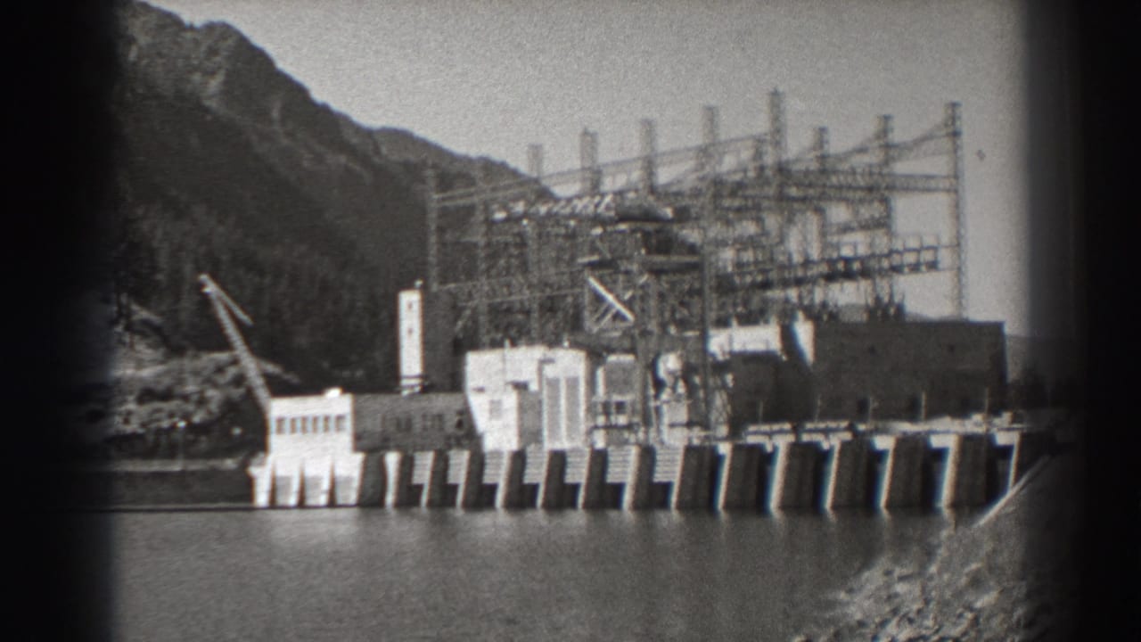 Dam buildings and electrical tower framework at the base of a wooded mountain slope.