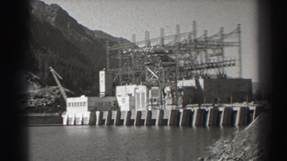 Archival 8mm frame of Bonneville Dam construction, showing a crane near concrete works and dense transmission structures against a mountain backdrop.