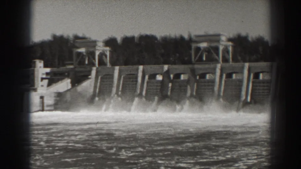 Black-and-white 8mm frame of Bonneville Dam, with water streaming from the spillway and a wooded ridge behind the structure.