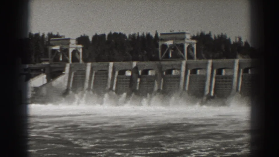 Wide 1938 home-movie view of Bonneville Dam in Oregon, showing the spillway, towers, river turbulence, and tree-covered background.