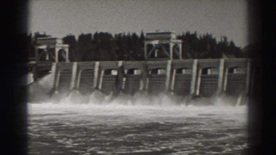 1938 8mm view of Bonneville Dam in Oregon, with water pouring through spillway bays and trees behind the concrete structure.