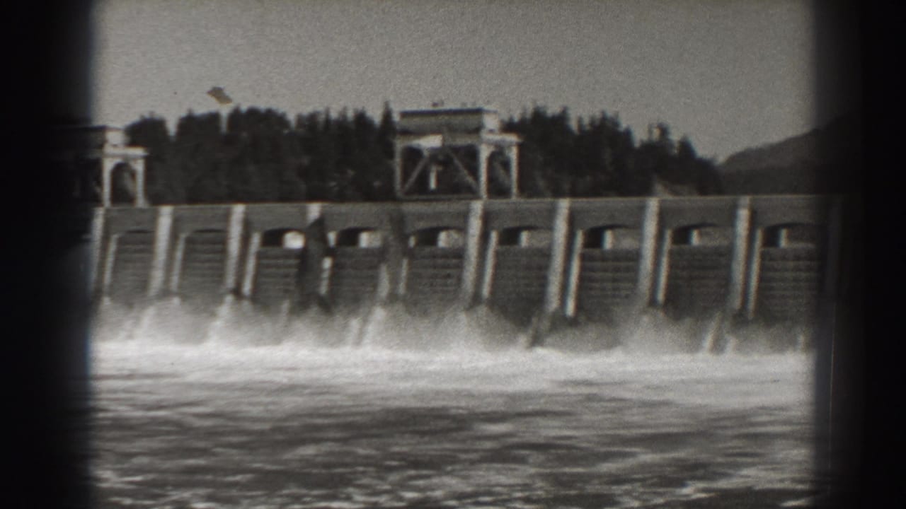 Dam spillway gates with white spray spreading across the river surface.