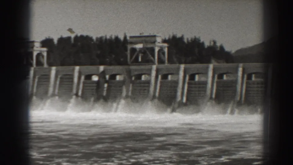 Bonneville Dam spillway flow filmed from across the water, with dark trees and a small gatehouse-like structure above the dam.