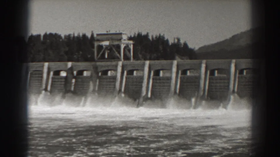 Black-and-white 8mm frame of Bonneville Dam, Oregon, showing repeated spillway bays and heavy white water at the base.