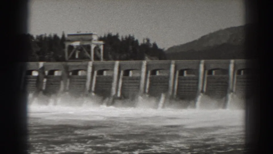 Late-1930s home-movie view of Bonneville Dam with spillways releasing water, wooded banks, and a hillside beyond the structure.