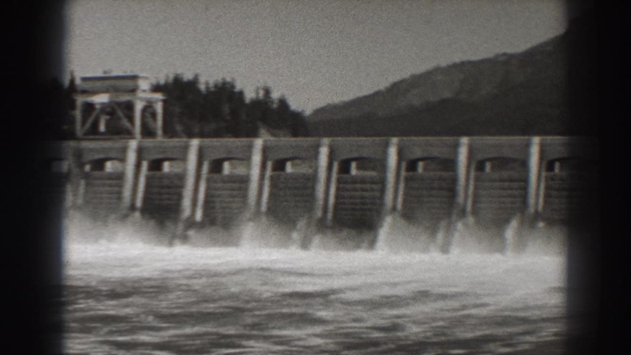 Concrete dam face with spillway spray and a small structure above.