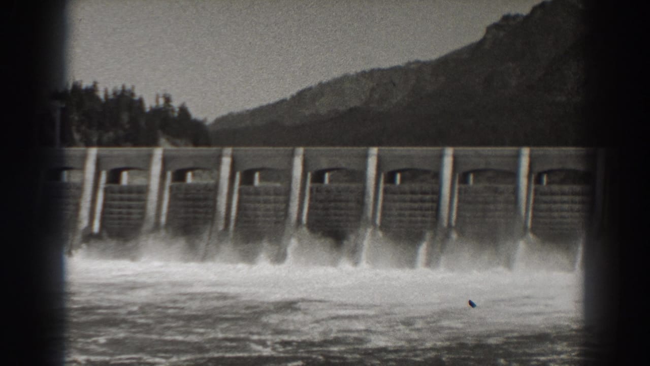 Concrete dam spillway releasing whitewater with mountains and trees behind it.