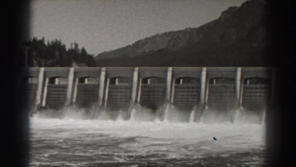 Bonneville Dam in Oregon, seen in 1938 8mm footage, with water spilling through concrete floodgates below a wooded mountain backdrop.