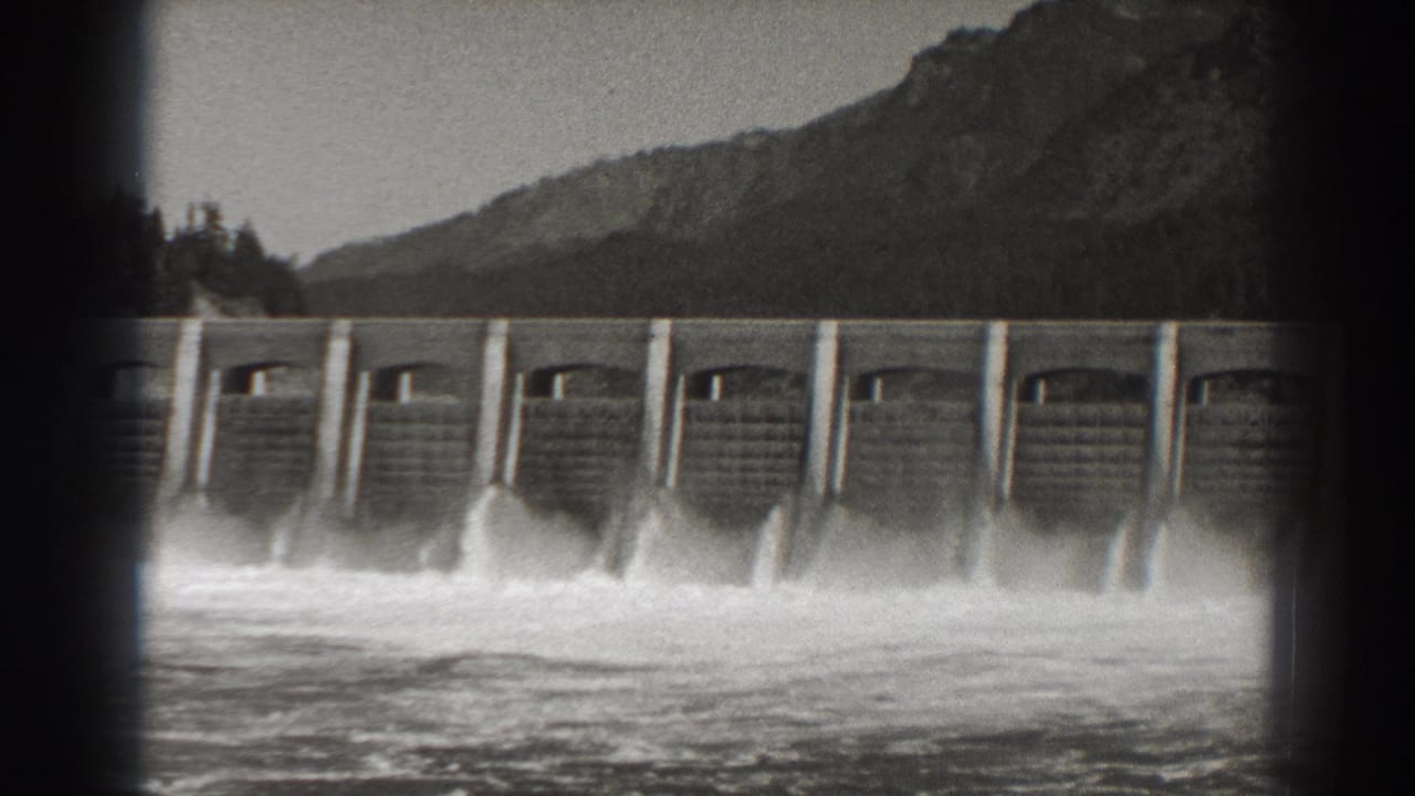 Dam floodgates release turbulent water with a dark mountain slope in the background.