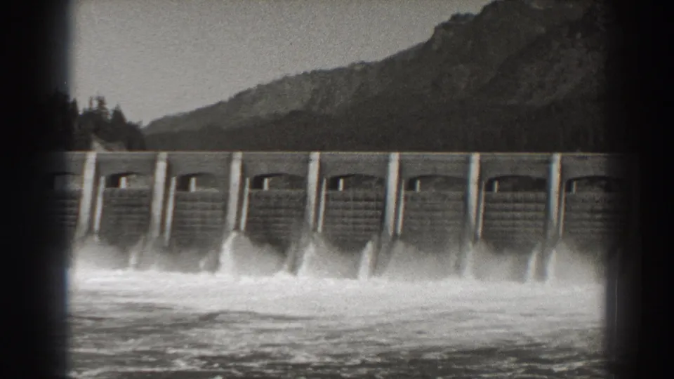 A 1938 view of Bonneville Dam shows several floodgate openings releasing churning water against forested slopes and a pale sky.