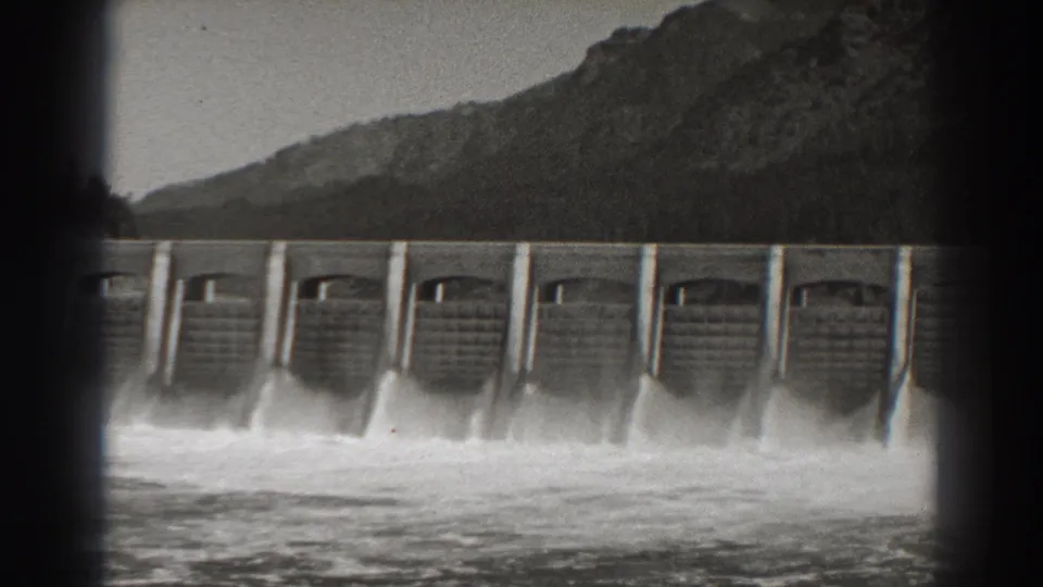 The frame holds on Bonneville Dam spillways, with whitewater spreading below the concrete structure and dark hills rising behind it.