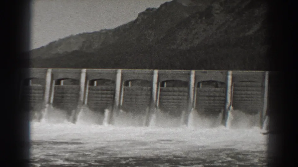 Bonneville Dam appears in a wide 1938 home-movie frame as water rushes from the spillway bays below a wooded mountain landscape.