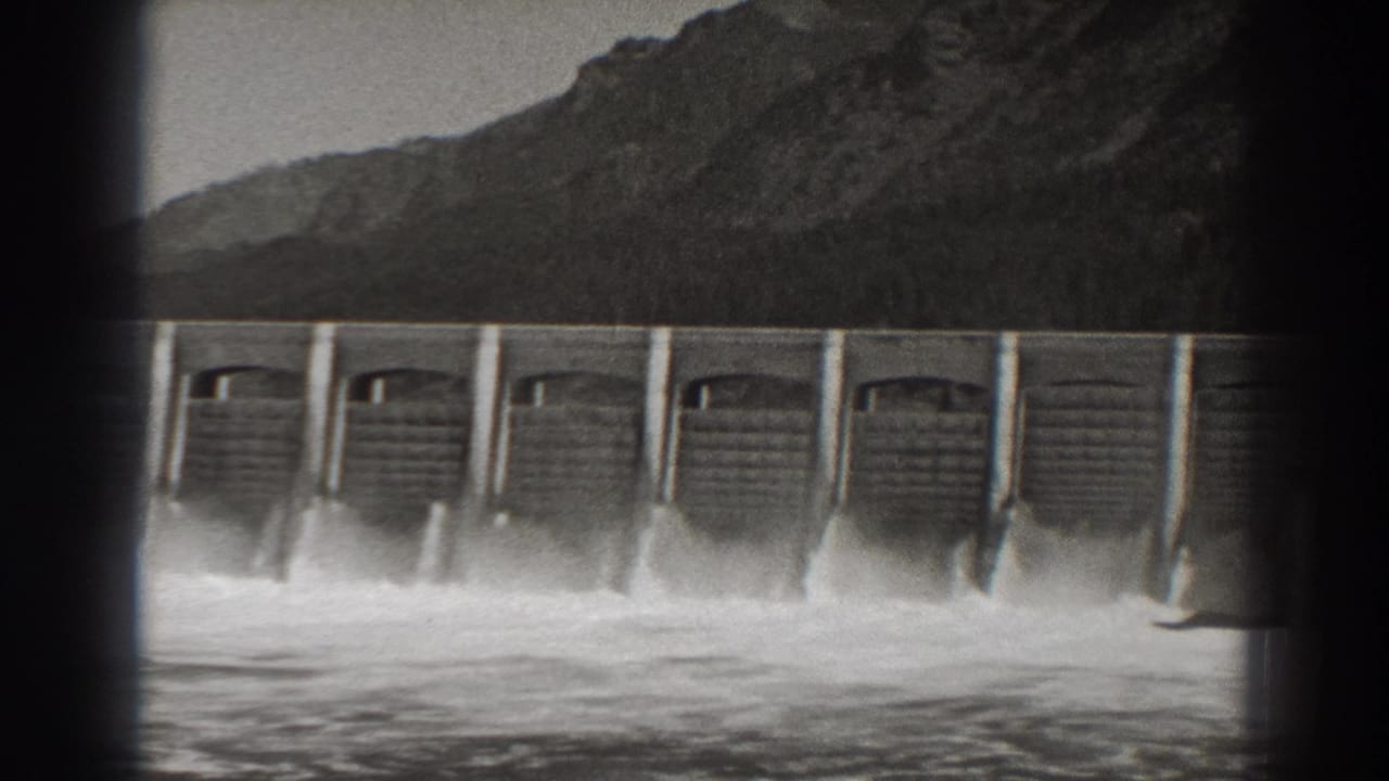 Concrete dam spilling white water with a mountain slope behind it.