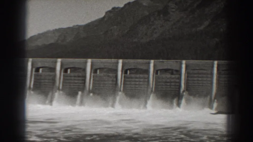 Bonneville Dam in Oregon releasing water through spillway bays, seen in black-and-white 1938 8mm footage with mountains behind.