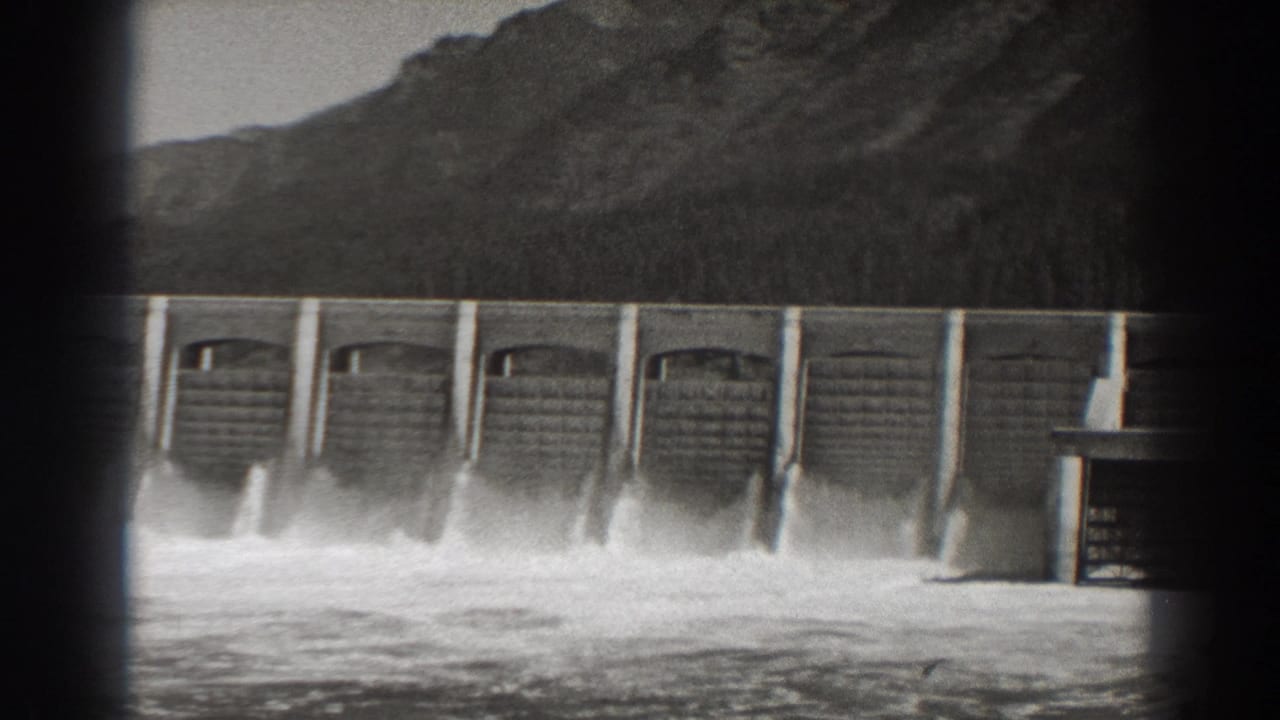 Wide view of dam spillways releasing water below a mountain ridge.