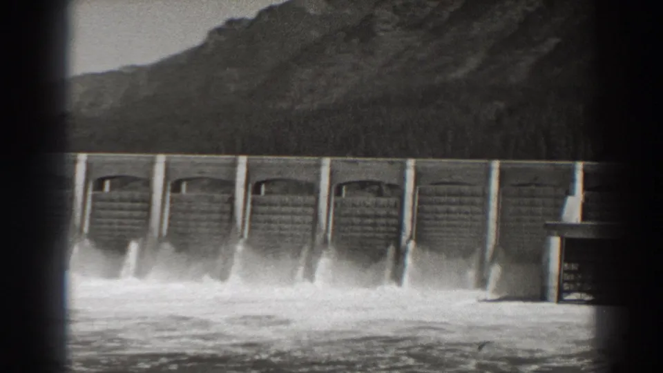 Black-and-white 8mm view of Bonneville Dam spillways releasing water, with a rugged Oregon mountain slope in the background.