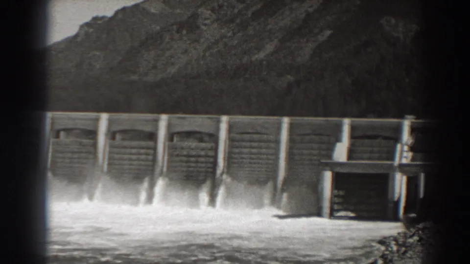 Bonneville Dam spillways discharge water in black-and-white 8mm footage, with mountain terrain behind and a small structure at right.