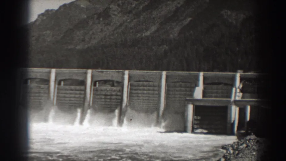 1938 8mm view of Bonneville Dam in Oregon, showing spillway gates releasing white water beneath wooded slopes.