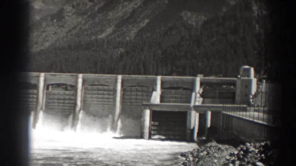 The pan across Bonneville Dam shows spillway bays, bright water discharge, a service structure, and rocky riverbank foreground.