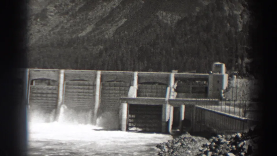 A 1938 home-movie frame of Bonneville Dam shows the right-side spillways, walkway railings, service building, and turbulent water.
