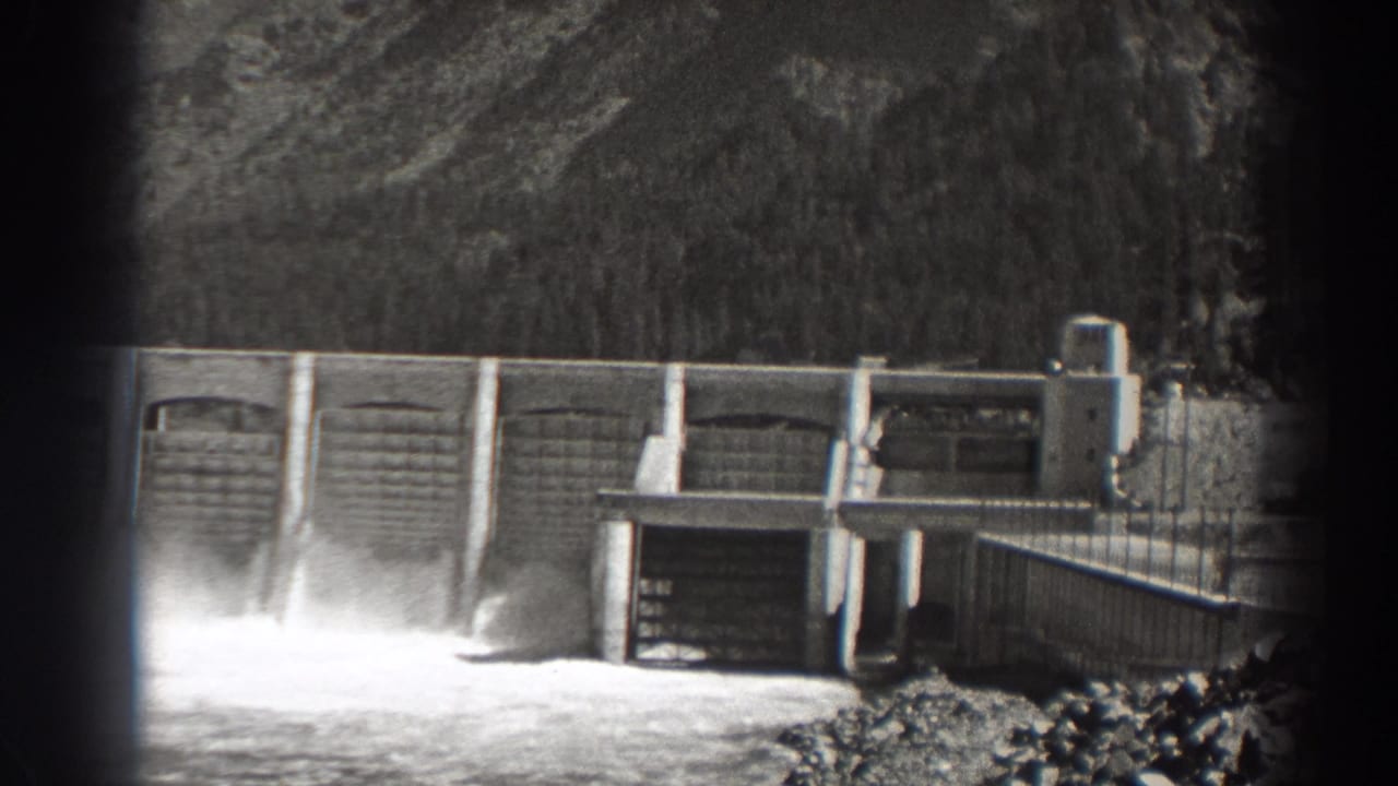 Spillway water surges below a concrete dam wall with wooded hills behind it.