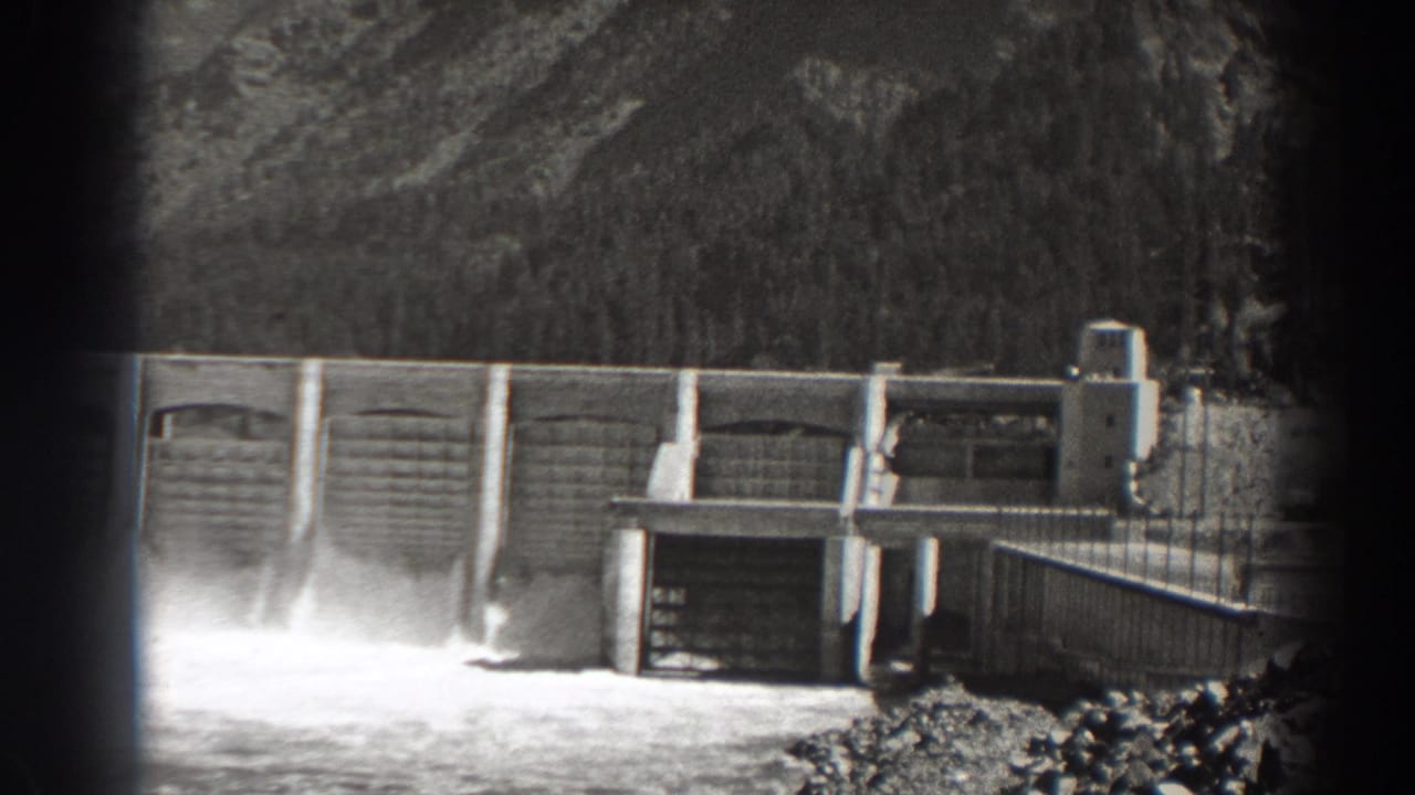 Concrete dam gates and a bright spillway plume appear below wooded terrain.