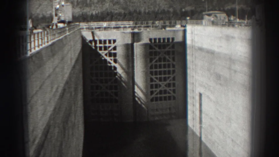 1938 Bonneville Dam view showing concrete walls, large gridded intake gates, railings, and heavy shadows over water.