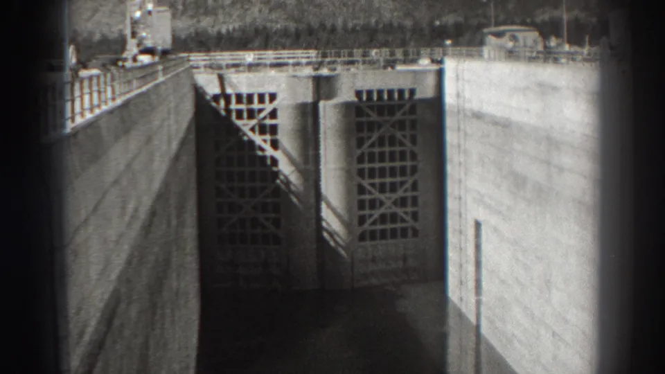 Bonneville Dam intake area in 1938, with gridded doors, concrete side walls, upper railings, and shadowed water below.