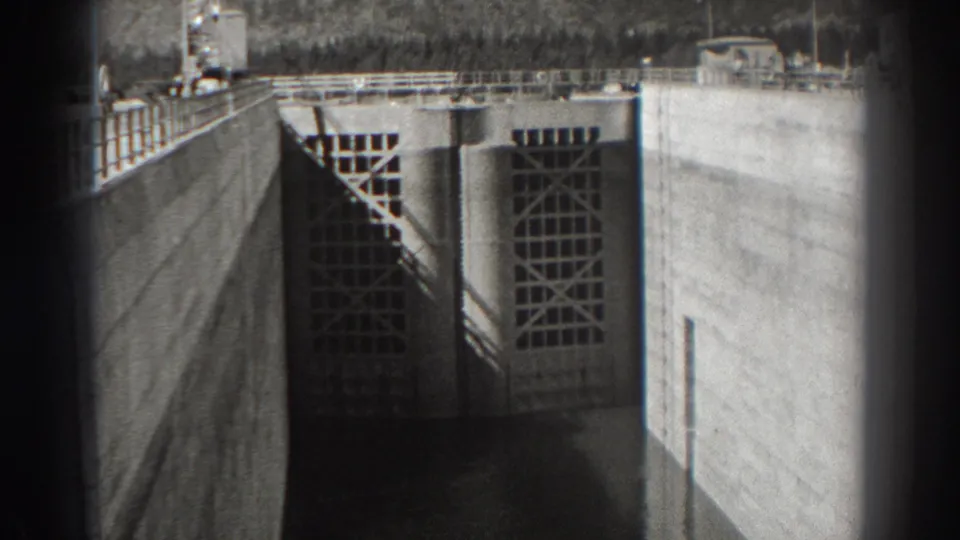 Black-and-white 8mm frame of Bonneville Dam's concrete intake gates, with railings above and dark water at the base.