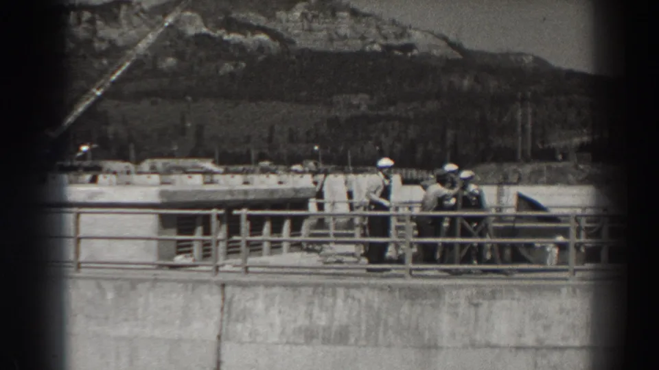 Workers in hard hats stand and talk on the Bonneville Dam upper walkway, with railings, concrete surfaces, and forested hills behind them.
