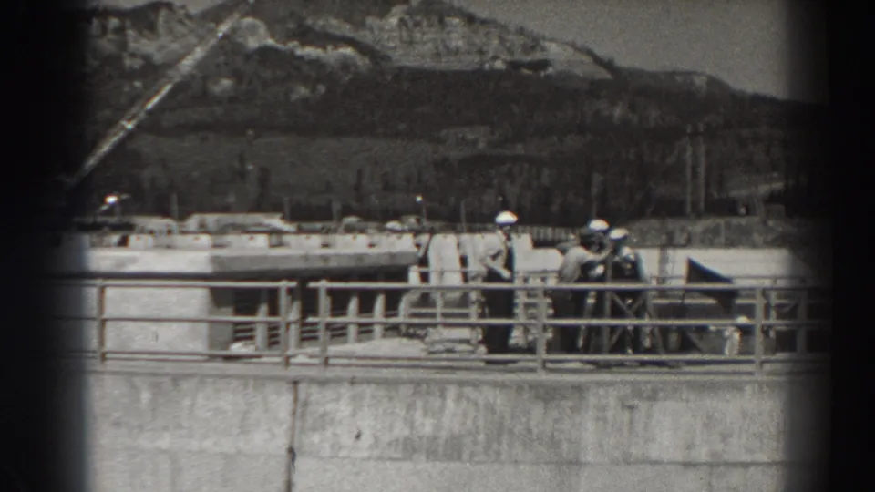 Bonneville Dam workers remain gathered on the upper walkway, framed by railings, concrete construction, distant hills, and black-and-white 8mm grain.