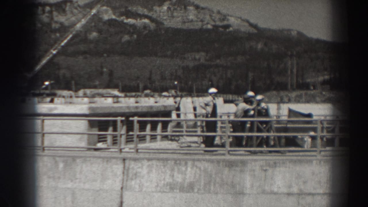 Men in hard hats stand behind a concrete railing at a dam construction site with hills behind them.